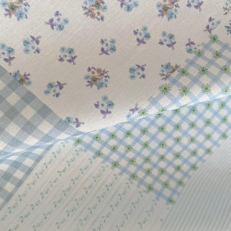 May include: Close-up of fabric with a floral pattern in shades of blue, purple, and tan on a white background. The fabric also features a light blue gingham and a light blue and white checkered pattern.