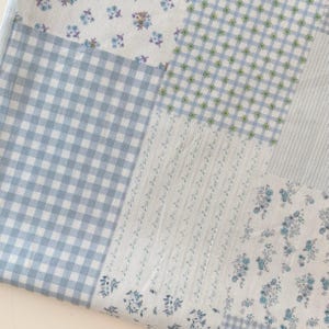 May include: A patchwork fabric with various patterns in shades of blue and white. The design includes gingham, floral, and striped sections, creating a vintage, cottagecore aesthetic. The fabric is likely cotton or a similar material.