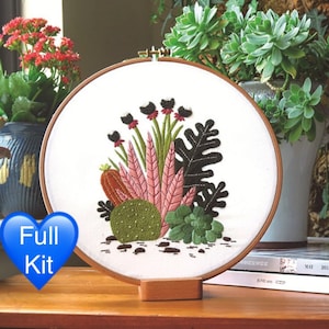 May include: A white embroidery hoop featuring a design of pink, green and black plants and flowers. The hoop is on a wooden stand with a book in the background.