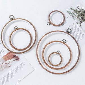 May include: Four embroidery hoops of different sizes, with a wooden frame and white fabric. The hoops are arranged in a staggered pattern on a white surface.