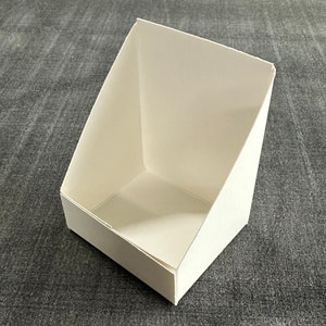 May include: A white cardboard display stand with a triangular shape and a rectangular opening. The stand is empty and ready to be filled with products.