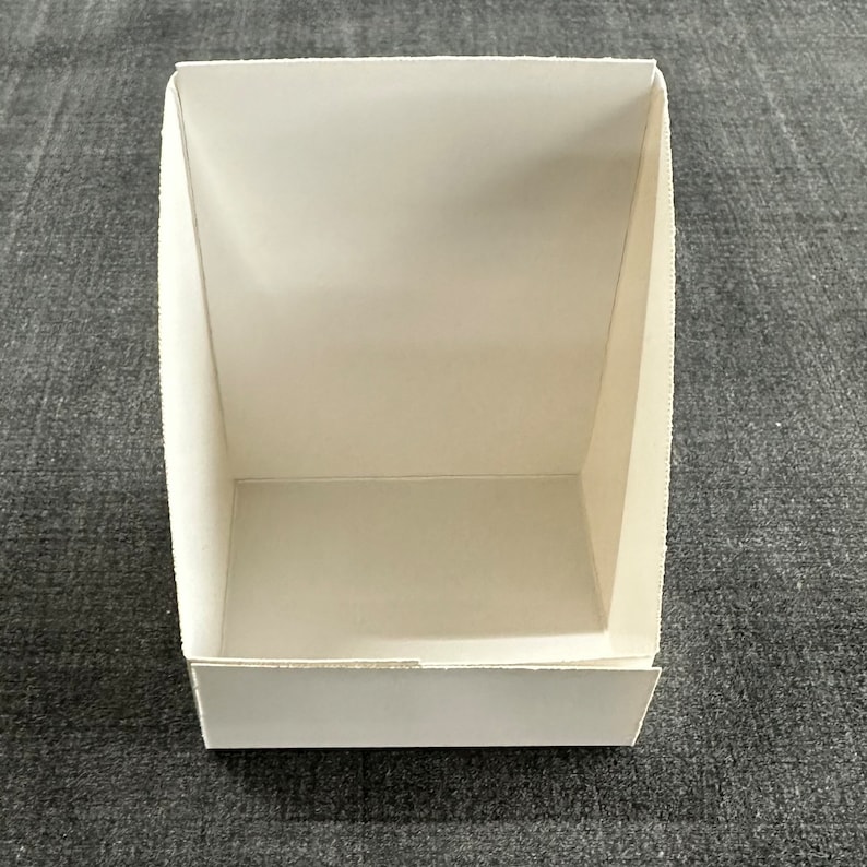 May include: A white cardboard box with a rectangular opening on the front. The box is empty and has a smooth surface.
