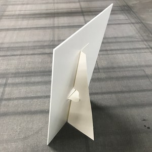 May include: White cardboard sign with a built-in easel stand. The stand is folded down and the sign is lying flat on a gray surface.