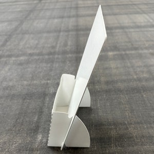 May include: White cardboard display stand with a slot for holding business cards or other small items. The stand has a vertical back panel and two angled feet for stability.
