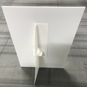 May include: White cardboard sign stand with a fold-out base. The stand is designed to hold a sign or poster.