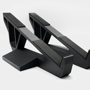 Pode incluir: Dois suportes pretos para laptop com plataformas angulares e almofadas de espuma. Os suportes têm um design geométrico com uma base e vigas de suporte. Eles são projetados para elevar um laptop para melhorar a ergonomia.