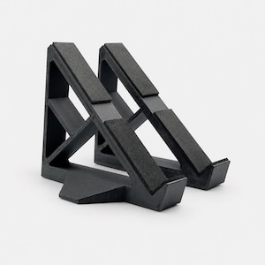 May include: A black triangular device stand featuring a rigid frame with two angled support arms. The structure includes textured grip pads along the top surfaces of the arms and a small base extension for stability on flat surfaces.
