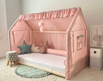 Puede incluir: Una estructura de cama de madera con forma de casa cubierta por un dosel de tela rosa con un borde superior con volantes. La cama incluye un colchón, un cojín en forma de estrella azul verdoso, uno de nube beige, uno de orejas de conejo y un rulo rosa.