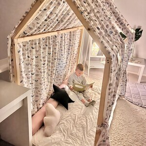 May include: A wooden playhouse fort with a white fabric canopy featuring a rainbow pattern. A child is sitting inside the fort reading a book.