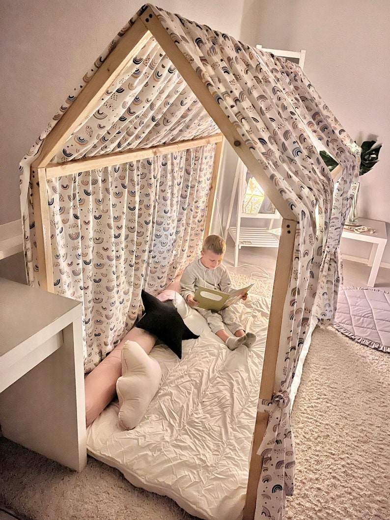 May include: A wooden playhouse fort with a white fabric canopy featuring a rainbow pattern. A child is sitting inside the fort reading a book.