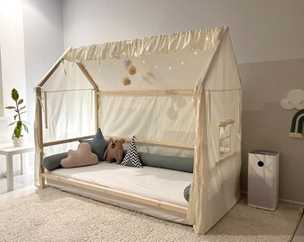 Könnte beinhalten: Ein hausförmiger Holzbettrahmen mit einem cremefarbenen Stoffhimmel mit gerüschtem Oberrand. Das Bett umfasst eine weiße Matratze, ein rosafarbenes wolkenförmiges Kissen, ein blaues sternförmiges Kissen, einen braunen Bären und einen schwarz-weißen Kegel.