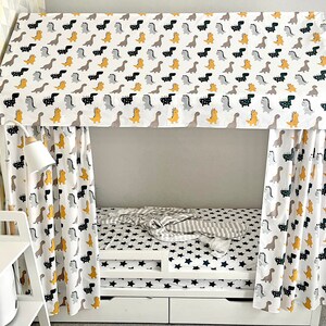May include: A white canopy bed with a dinosaur print fabric. The bed has a white mattress with a gray and white striped blanket and a black star patterned sheet. The bed has a white wooden frame and two white drawers.