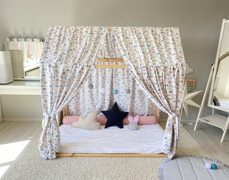 May include: A white canopy bed with rainbow patterned curtains and a white mattress. The bed has a white, pink, and blue pillow on it. The bed is in a room with a white rug and a gray rug with a white border.