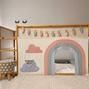 Puede incluir: Una cama alta de madera con un panel de tela con tem&aacute;tica de arco&iacute;ris. El panel presenta un arco de arco&iacute;ris, una nube rosa y una nube azul con texto. Un peque&ntilde;o taburete con un patr&oacute;n de estrellas est&aacute; cerca. Elementos decorativos incluyen una guirnalda y arte mural.