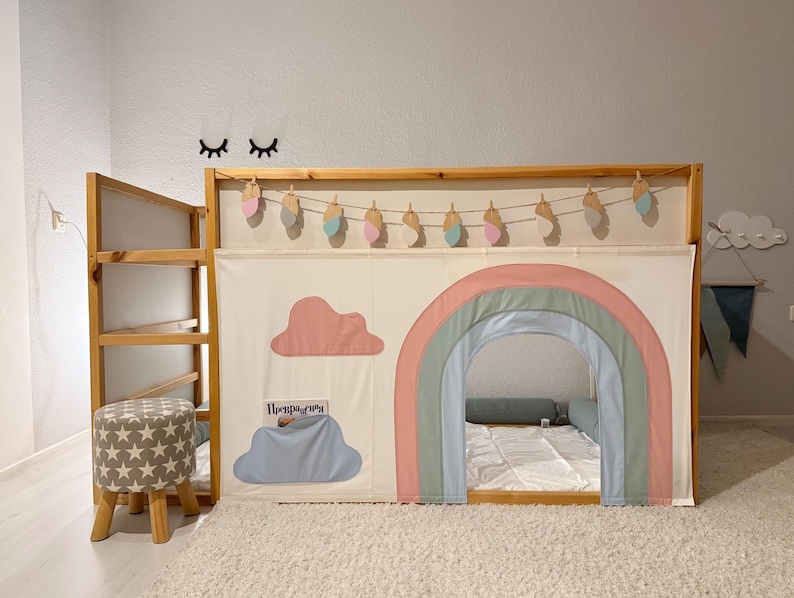 Puede incluir: Una cama alta de madera con un panel de tela con tem&aacute;tica de arco&iacute;ris. El panel presenta un arco de arco&iacute;ris, una nube rosa y una nube azul con texto. Un peque&ntilde;o taburete con un patr&oacute;n de estrellas est&aacute; cerca. Elementos decorativos incluyen una guirnalda y arte mural.