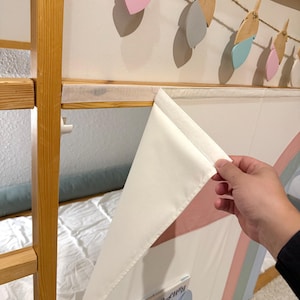 Puede incluir: Una litera de madera con un panel de tela blanco y de colores pastel. El panel tiene un bolsillo con un libro dentro. Encima de la cama, cuelga una guirnalda de plumas de madera y de colores pastel.