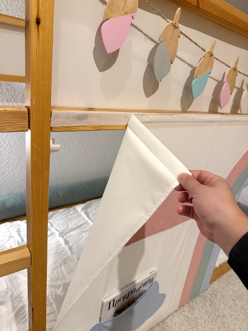Puede incluir: Una litera de madera con un panel de tela blanco y de colores pastel. El panel tiene un bolsillo con un libro dentro. Encima de la cama, cuelga una guirnalda de plumas de madera y de colores pastel.