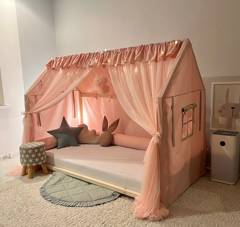 Op de afbeelding: Een huisvormig bedframe met een lichtroze stoffen hoes, voorzien van doorschijnende tule gordijnen, een gerimpelde bovenrand en ge&iuml;ntegreerde lichtsnoeren. Het interieur bevat een grijs stervormig kussen, een wolk-vormig kussen en een bruin konijnkussen.