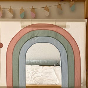 Puede incluir: Un marco de cama de madera con un arco de arco&iacute;ris de tela. El arco&iacute;ris presenta tonos apagados de rosa, verde y azul. Una guirnalda de decoraciones de plumas de madera cuelga sobre el arco. La cama est&aacute; cubierta con un edred&oacute;n blanco.