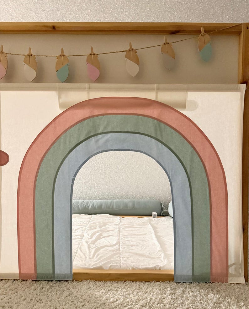 Puede incluir: Un marco de cama de madera con un arco de arco&iacute;ris de tela. El arco&iacute;ris presenta tonos apagados de rosa, verde y azul. Una guirnalda de decoraciones de plumas de madera cuelga sobre el arco. La cama est&aacute; cubierta con un edred&oacute;n blanco.