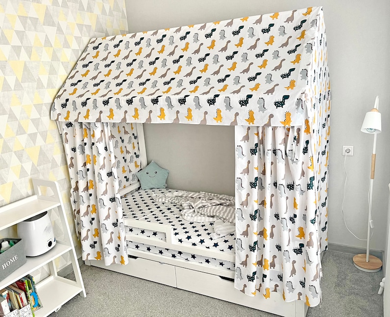 May include: White canopy bed with a dinosaur print fabric. The bed has a white wooden frame and a white mattress with a black and white star pattern. The canopy is made of a white fabric with a yellow and black dinosaur print.