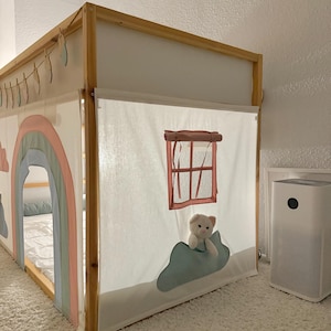 Puede incluir: Cama infantil con estructura de madera y dosel de tela. El dosel est&aacute; decorado con un motivo de arco&iacute;ris y nubes. Un peque&ntilde;o osito de peluche est&aacute; colocado cerca de una ventana. La cama est&aacute; sobre una alfombra blanca.