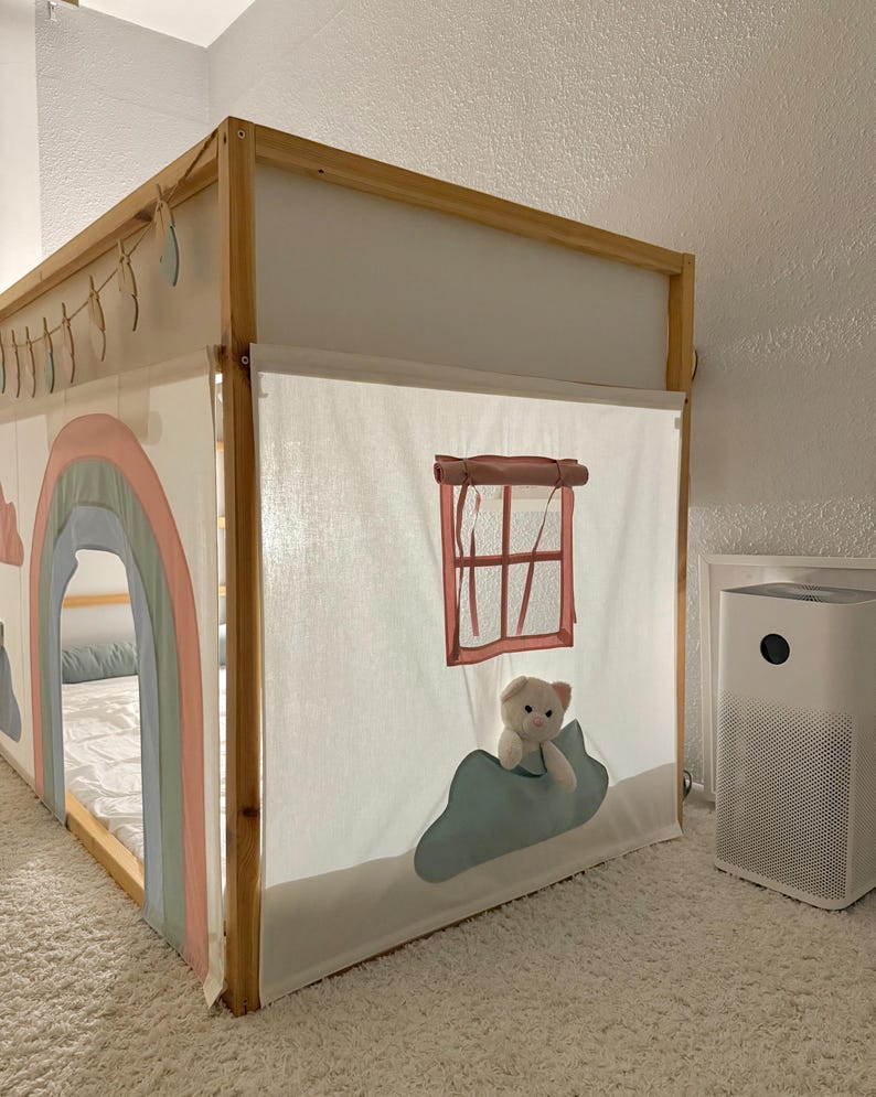 Puede incluir: Cama infantil con estructura de madera y dosel de tela. El dosel est&aacute; decorado con un motivo de arco&iacute;ris y nubes. Un peque&ntilde;o osito de peluche est&aacute; colocado cerca de una ventana. La cama est&aacute; sobre una alfombra blanca.