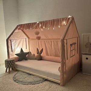 Op de afbeelding: Een huisvormig bedframe met een lichtroze stoffen hoes, voorzien van een gerimpelde bovenrand met ge&iuml;ntegreerde lichtsnoeren. Het bed bevat een wit matras, een lang roze rolkussen en decoratieve kussens in de vorm van een ster, wolk en konijn.