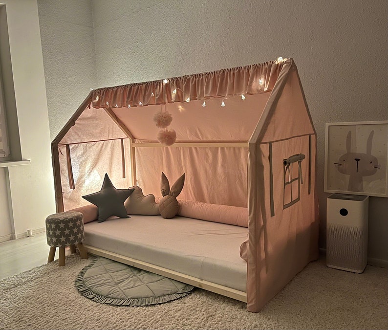 Op de afbeelding: Een huisvormig bedframe met een lichtroze stoffen hoes, voorzien van een gerimpelde bovenrand met ge&iuml;ntegreerde lichtsnoeren. Het bed bevat een wit matras, een lang roze rolkussen en decoratieve kussens in de vorm van een ster, wolk en konijn.