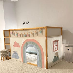 Puede incluir: Una cama alta de madera con un cerramiento de tela con tem&aacute;tica de arco&iacute;ris. El cerramiento presenta un arco&iacute;ris pastel, nubes y una ventana con un osito de peluche. Tambi&eacute;n se ven una peque&ntilde;a escalera de madera y una guirnalda decorativa.