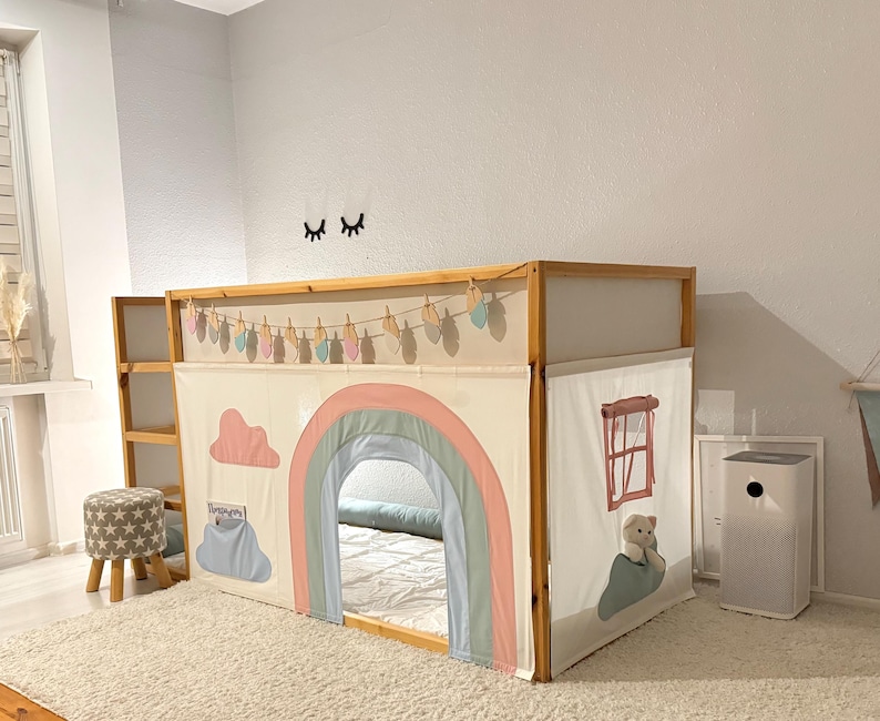 Puede incluir: Una cama alta de madera con un cerramiento de tela con tem&aacute;tica de arco&iacute;ris. El cerramiento presenta un arco&iacute;ris pastel, nubes y una ventana con un osito de peluche. Tambi&eacute;n se ven una peque&ntilde;a escalera de madera y una guirnalda decorativa.