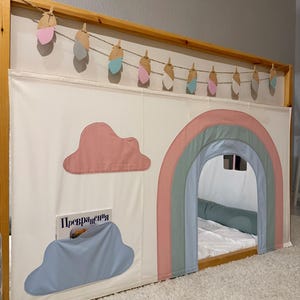 Puede incluir: Un marco de cama de madera con un dosel de tela blanca con un arco iris y aplicaciones de nubes. Una guirnalda de recortes decorativos en forma de pluma cuelga por encima. Un libro es visible en un bolsillo en forma de nube.