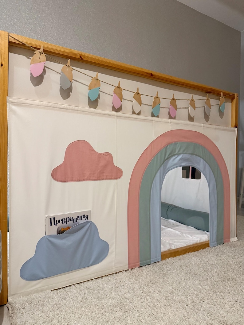 Puede incluir: Un marco de cama de madera con un dosel de tela blanca con un arco iris y aplicaciones de nubes. Una guirnalda de recortes decorativos en forma de pluma cuelga por encima. Un libro es visible en un bolsillo en forma de nube.