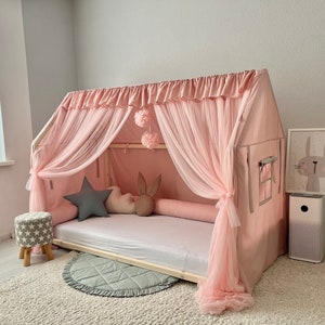Op de afbeelding: Een houten bedframe in de vorm van een huis, gedrapeerd met lichtroze stof en transparante tule gordijnen. Het bed bevat een wit matras, een grijs stervormig kussen, een wit wolk-vormig kussen en een bruin kussen met konijnenoren.
