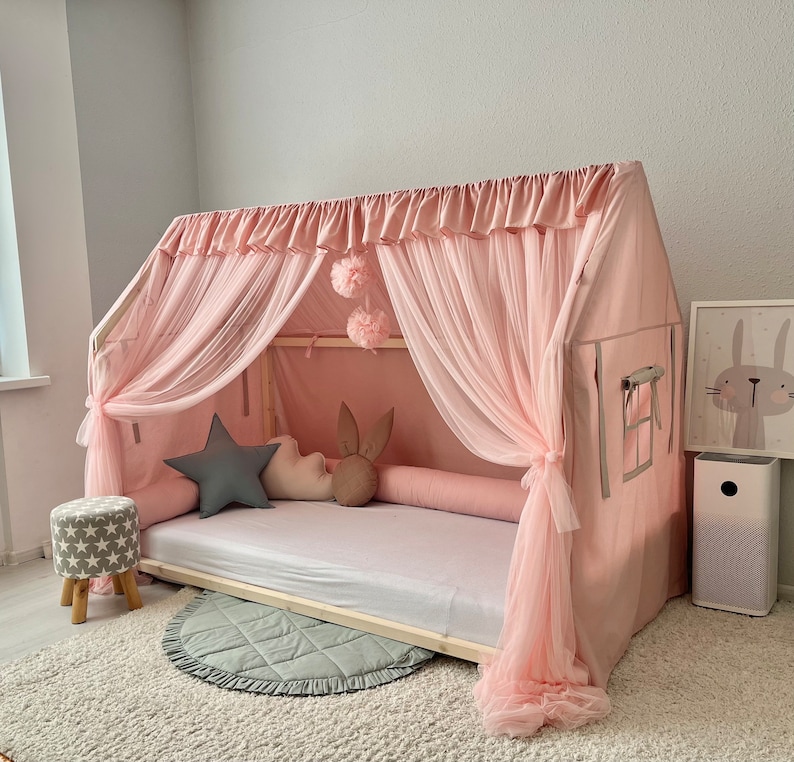 Op de afbeelding: Een houten bedframe in de vorm van een huis, gedrapeerd met lichtroze stof en transparante tule gordijnen. Het bed bevat een wit matras, een grijs stervormig kussen, een wit wolk-vormig kussen en een bruin kussen met konijnenoren.