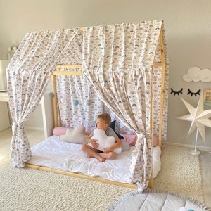 May include: A wooden bed frame with a white canopy featuring a rainbow pattern. The bed is made up with white bedding and a pink and white pillow. A child is sitting on the bed holding a white cloud-shaped pillow. The text "Dream Big" is visible on a wooden sign.