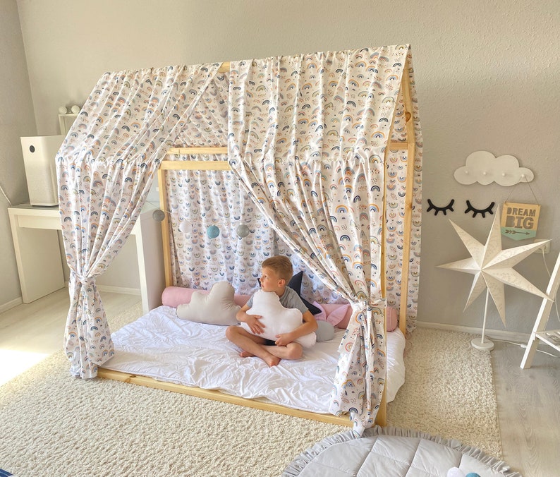 May include: A wooden bed frame with a white canopy featuring a rainbow pattern. The bed is made up with white bedding and a pink and white pillow. A child is sitting on the bed holding a white cloud-shaped pillow. The text "Dream Big" is visible on a wooden sign.