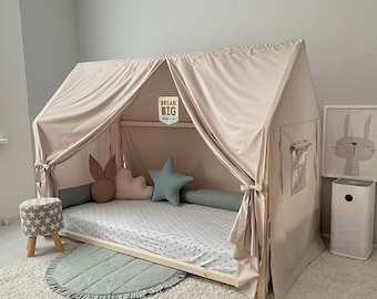 May include: A wooden house-shaped bed frame draped with a beige fabric canopy. The bed features a white sheet with gray stars, a sage green bolster pillow, a star-shaped pillow, a cloud-shaped pillow, and a brown bunny-ear pillow. A sign reads DREAM BIG.