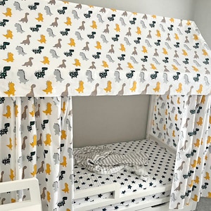 May include: A white bed canopy with a dinosaur print. The canopy is made of fabric and has a yellow, green, and gray dinosaur pattern. The bed is white and has a gray and white striped blanket on it.