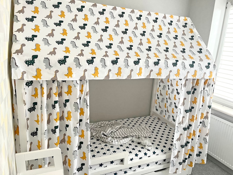 May include: A white bed canopy with a dinosaur print. The canopy is made of fabric and has a yellow, green, and gray dinosaur pattern. The bed is white and has a gray and white striped blanket on it.
