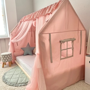 Op de afbeelding: Een huisvormig bedframe bedekt met een lichtroze stoffen overkapping met een geplooide bovenrand en doorschijnende gordijnen. De structuur bevat een zijraam met opbindbare stoffen luiken, een wit matras en een grijs stervormig kussen.
