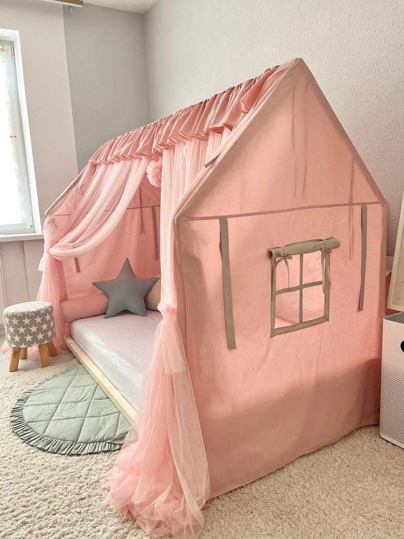 Op de afbeelding: Een huisvormig bedframe bedekt met een lichtroze stoffen overkapping met een geplooide bovenrand en doorschijnende gordijnen. De structuur bevat een zijraam met opbindbare stoffen luiken, een wit matras en een grijs stervormig kussen.