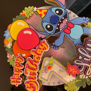 Pu&ograve; includere: Un topper per torta blu con il personaggio di Stitch con un papillon rosso, circondato da palloncini colorati, fiori e le parole "Happy Birthday" e "Lilly".