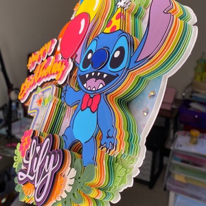 Pu&ograve; includere: Un topper per torta a pi&ugrave; livelli con un personaggio Stitch blu di Lilo & Stitch, che indossa un cappello da festa e tiene un palloncino rosso. Il topper &egrave; decorato con strisce arcobaleno, foglie verdi e fiori. Il testo "Happy Birthday Lily" &egrave; scritto in viola e bianco sullo strato inferiore.