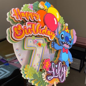 Pu&ograve; includere: Un topper per torta colorato con un personaggio Stitch blu di Lilo & Stitch, con un cappello da festa, un papillon rosso e una mano alzata. Il topper include anche un numero 7 a strati, le parole "Happy Birthday" e "Lily", e fiori e foglie tropicali.