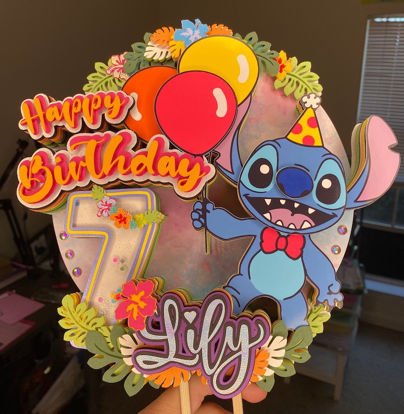 Pu&ograve; includere: Un topper per torta colorato con Stitch di Lilo & Stitch. Il topper include un Stitch blu che tiene palloncini, un numero 7, le parole "Happy Birthday" e "Lily", e foglie e fiori tropicali.