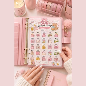 May include: A pink six-ring binder containing a 500 Savings Challenge tracker sheet. The sheet features cartoon illustrations of cats and bears, with a grid of icons representing various dollar amounts, including $5, $10, $15, $20, $25, $30, $35, $40, and $50.