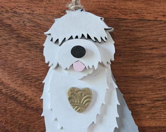 May include: A layered wooden ornament shaped like a shaggy dog with white fur, a black nose, a pink tongue, and a gold heart-shaped emblem on its chest. The dog figure is suspended by a natural jute twine loop attached to the top of its head.