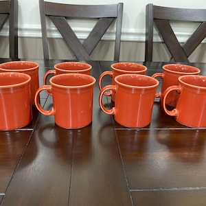 Op de afbeelding: Een set van acht oranje keramische mokken met afgeronde handvatten. De mokken staan op een donkere houten tafel, met een paar houten stoelen op de achtergrond. De mokken hebben een klassiek ontwerp.