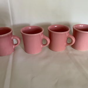 Puede incluir: Cuatro tazas rosas de cerámica con asas dispuestas en fila. Las tazas tienen forma redondeada y acabado brillante. Las tazas son de color rosa claro liso.